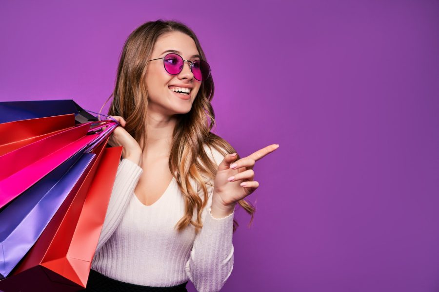 beautiful-smiling-young-blonde-woman-pointing-sunglasses-holding-shopping-bags-credit-card-pink-wall